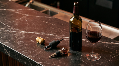 Gold champagne stopper, black pour-spout stopper, and glass bottle stopper arranged next to an open wine bottle and glass on a dark marble counter