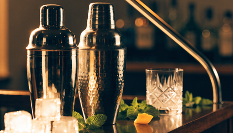 Stainless steel Boston and cobbler cocktail shakers with a crystal rocks glass, mint, and citrus on a dark wood home bar cart