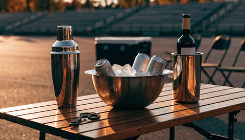 Styled tailgate bar setup on a folding table with a cocktail shaker, ice bucket, and bottle chiller for game day