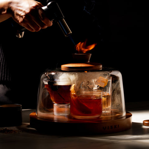 Smoke infusing under glass dome over whiskey cocktails — Viski Alchemi smoked cocktail kit in use